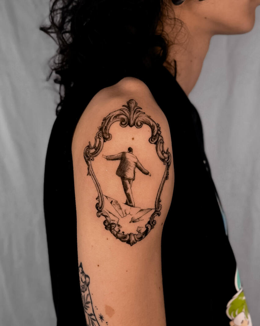 Tatuaggio fine line di una figura in equilibrio su un vetro all’interno di una cornice vintage ornamentale, con dettagli realistici e ombreggiature soft sul braccio